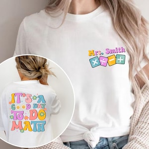 May include: A white t-shirt featuring a small graphic on the chest with the text Mrs. Smith above colorful math symbols. The back of the shirt displays the text IT'S A GOOD DAY TO DO MATH in large, colorful, retro-style letters with flower accents.