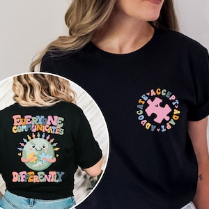 May include: Black t-shirt with a graphic of a smiling earth holding a puzzle piece. The text "Everyone Communicates Differently" is printed in rainbow colors. The back of the shirt has a pink puzzle piece in a circle with the text "Advocate Accept Adapt" in rainbow colors.