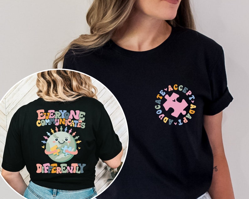 May include: Black t-shirt with a graphic of a smiling earth holding a puzzle piece. The text "Everyone Communicates Differently" is printed in rainbow colors. The back of the shirt has a pink puzzle piece in a circle with the text "Advocate Accept Adapt" in rainbow colors.