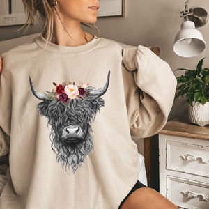 Puede incluir: Una sudadera beige con una ilustraci&oacute;n en blanco y negro de una vaca de las tierras altas con una corona de flores. Las flores son rosas, rojas y blancas.