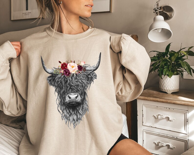 Puede incluir: Una sudadera beige con una ilustraci&oacute;n en blanco y negro de una vaca de las tierras altas con una corona de flores. Las flores son rosas, rojas y blancas.
