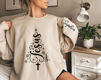May include: Beige sweatshirt with a black Christmas tree design. The tree is formed by the word "Jesus" and decorated with snowflakes, stars, and a cross. The sleeve has the name "Sophia" in black script.
