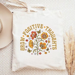 May include: A white canvas tote bag with a floral design and the text "Grow Positive Thoughts" in gold lettering.