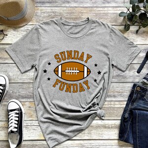 Pode incluir: Camiseta cinza com um gr&aacute;fico de futebol americano marrom e o texto "SUNDAY FUNDAY" em laranja.