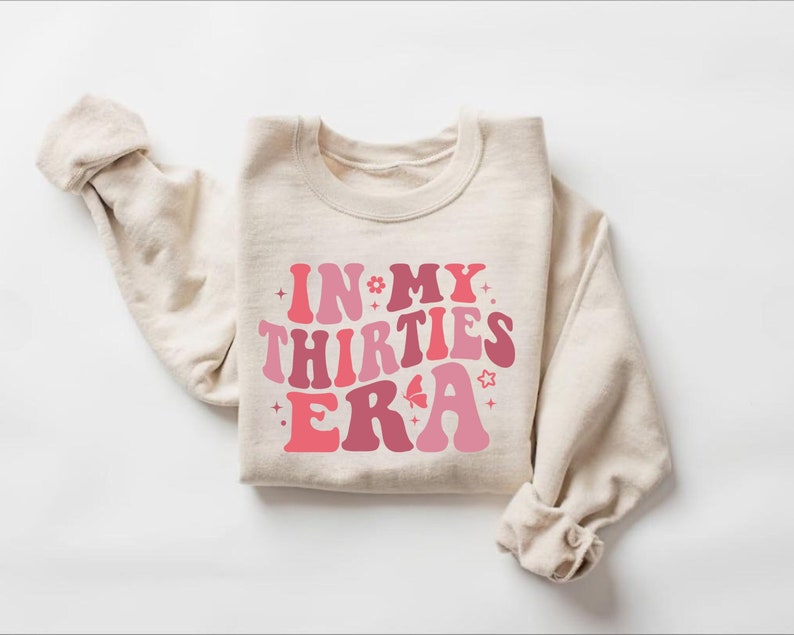 May include: A beige sweatshirt with the text "IN MY THIRTIES ERA" in pink and red retro font with a butterfly and stars.