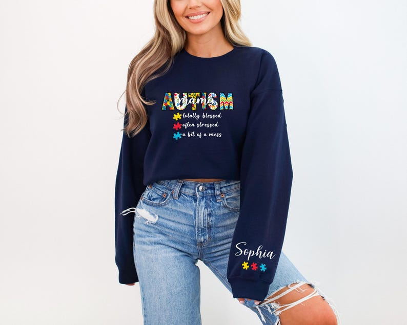 May include: A navy blue cropped sweatshirt with the text "Autism" in colorful puzzle pieces and the text "totally blessed, often stressed, a bit of a mess". The sweatshirt has a white personalized name "Sophia" on the sleeve with colorful puzzle pieces.