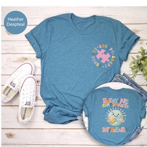 May include: Two blue t-shirts with a graphic design. The front shirt has a pink puzzle piece inside a circle with the text "PT-ADAPTA" and "EDUCATE". The back shirt has a cartoon earth with a puzzle piece and the text "EVERY NE COMMUNICATES DIFFERENTL".