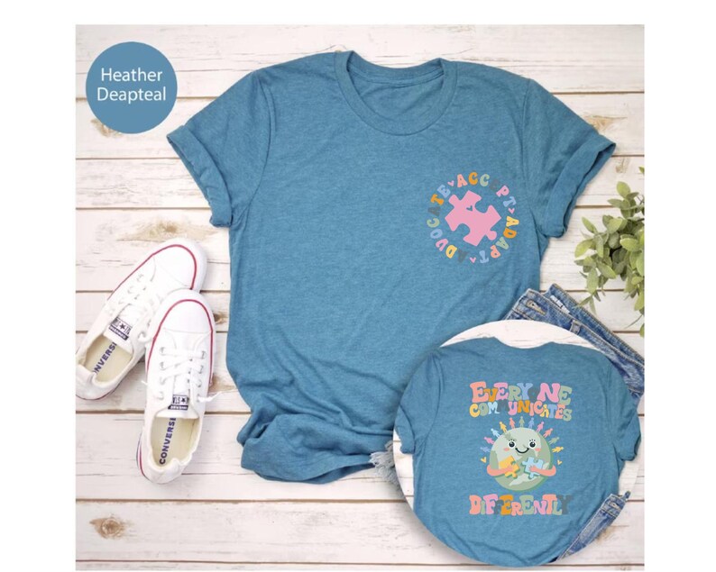 May include: Two blue t-shirts with a graphic design. The front shirt has a pink puzzle piece inside a circle with the text "PT-ADAPTA" and "EDUCATE". The back shirt has a cartoon earth with a puzzle piece and the text "EVERY NE COMMUNICATES DIFFERENTL".