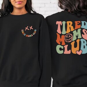 Puede incluir: Sudadera negra con un gr&aacute;fico colorido de "Tired Moms Club" en la espalda y una versi&oacute;n m&aacute;s peque&ntilde;a del gr&aacute;fico en la parte delantera. El gr&aacute;fico presenta una cara sonriente con X para los ojos.