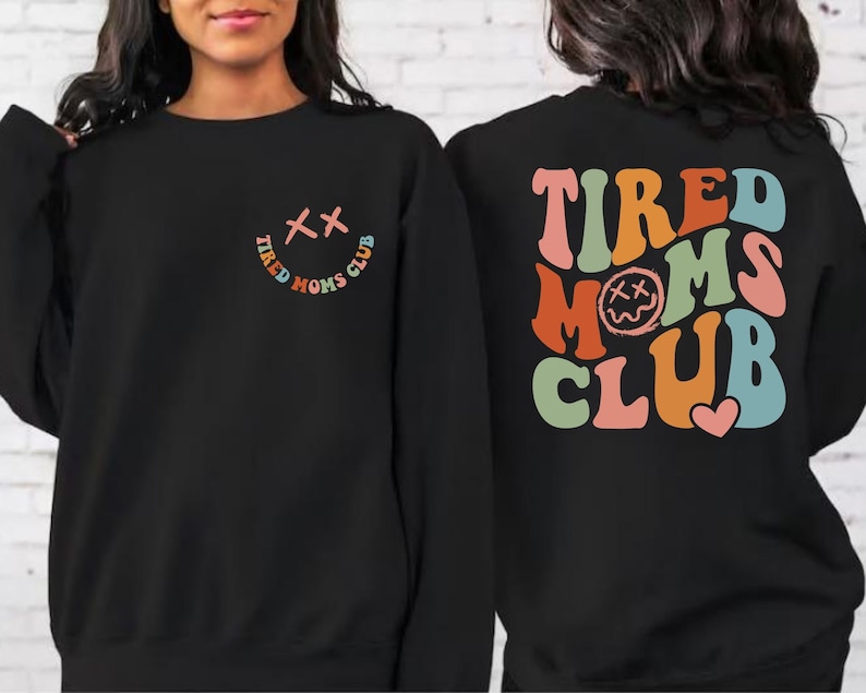 Puede incluir: Sudadera negra con un gr&aacute;fico colorido de "Tired Moms Club" en la espalda y una versi&oacute;n m&aacute;s peque&ntilde;a del gr&aacute;fico en la parte delantera. El gr&aacute;fico presenta una cara sonriente con X para los ojos.