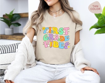 May include: A tan short-sleeved t-shirt featuring the text "FIRST GRADE VIBES" in a colorful, rounded font. The design includes small flower and star graphics, along with a pencil icon, all rendered in shades of pink, blue, yellow, and green.
