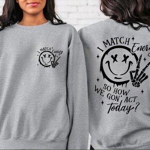 Puede incluir: Sudadera gris con un estampado gr&aacute;fico negro en la parte delantera y trasera. El gr&aacute;fico presenta una cara sonriente con X para los ojos, un efecto de goteo y una mano de esqueleto haciendo un signo de paz. El texto dice "I Match Energy So How We Gon' Act Today?"