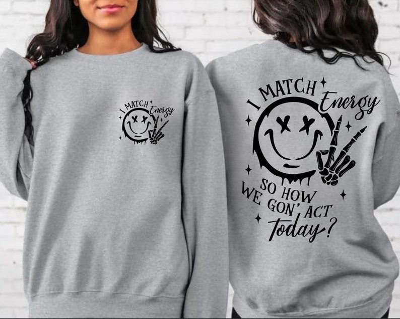 Puede incluir: Sudadera gris con un estampado gr&aacute;fico negro en la parte delantera y trasera. El gr&aacute;fico presenta una cara sonriente con X para los ojos, un efecto de goteo y una mano de esqueleto haciendo un signo de paz. El texto dice "I Match Energy So How We Gon' Act Today?"
