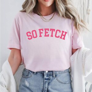 Op de afbeelding: Roze T-shirt met de tekst "SO FETCH" in een roze, universitaire stijl lettertype.