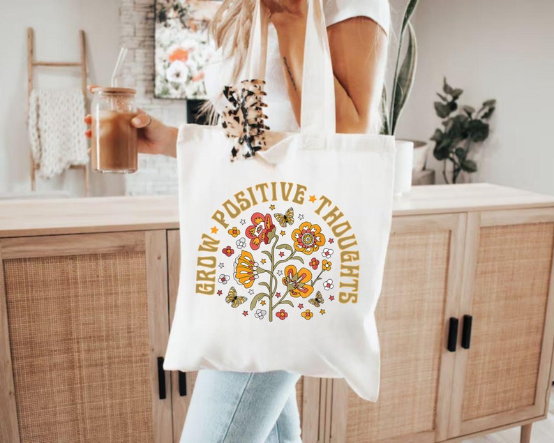May include: White canvas tote bag with a floral design and the text "Grow Positive Thoughts" in a retro font.