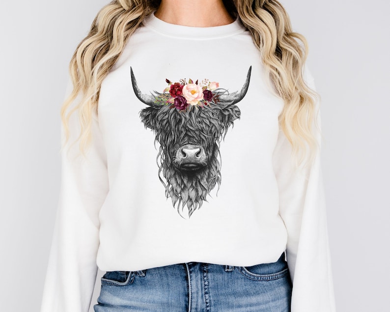 Puede incluir: Sudadera blanca con una ilustraci&oacute;n en blanco y negro de una vaca de las tierras altas con una corona floral de flores burdeos y rosas.