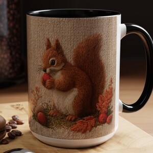 May include: A white ceramic mug with a black handle and interior, featuring a detailed illustration of a brown squirrel holding a red nut, surrounded by autumn leaves and berries on a textured beige background that mimics a woven fabric surface.