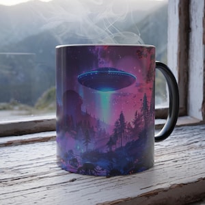 May include: A black and white ceramic mug featuring a colorful illustration of a UFO hovering over a forest at night. The design includes purple, pink, and blue hues, with a beam of light shining down. The mug has a black handle and a white interior.