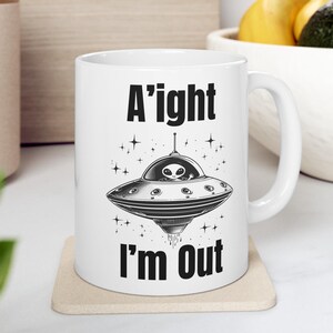 May include: White ceramic mug with a black and white illustration of a UFO with an alien inside. The text "A'ight I'm Out" is printed on the mug.