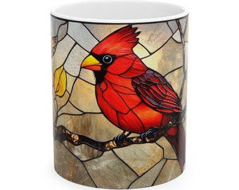 May include: A white ceramic mug with a stained glass design of a red cardinal perched on a branch. The cardinal has a yellow beak and black eyes. The background is a mosaic of brown, yellow, and gray.
