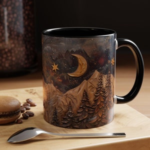 May include: A black ceramic mug with a mountain scene and a crescent moon with stars. The mug has a wooden texture and a black handle.