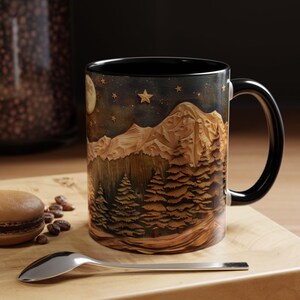 May include: A black ceramic mug with a mountain scene and a starry night sky. The mug features a detailed illustration of a mountain range with trees in the foreground. The sky is dark blue with white stars and a crescent moon.