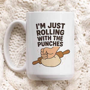 May include: White ceramic mug with a handle featuring the text "I'M JUST ROLLING WITH THE PUNCHES" in brown letters. The mug has a cartoon illustration of a hand rolling dough with a rolling pin. The mug is on a white knitted surface.