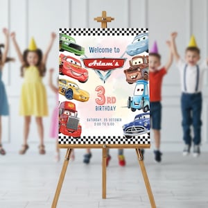 May include: A wooden easel displays a colorful birthday sign featuring animated car characters. The sign reads "Welcome to Adam's 3rd Birthday" with the date and time. The background is a watercolor wash with checkered borders.