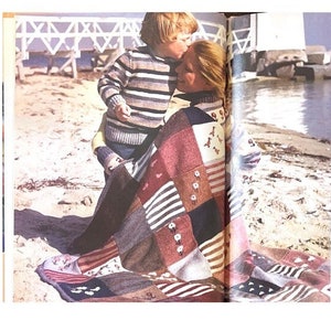 May include: A woman and child are sitting on a beach, wrapped in a colourful patchwork blanket. The blanket is made of different coloured squares with various patterns, including stripes, flowers, and geometric designs.