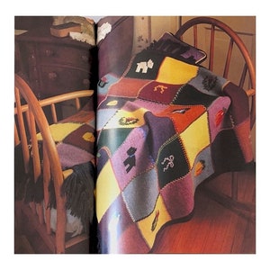 Puede incluir: Una manta de bebé de punto de colores con un diseño de patchwork que presenta varios motivos de animales y festivos. La manta está drapeada sobre una silla de madera.