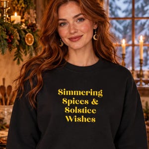 May include: A black crewneck sweatshirt featuring the yellow text Simmering Spices & Solstice Wishes printed on the front. The garment has long sleeves and a ribbed neckline, providing a simple and functional design for casual wear.