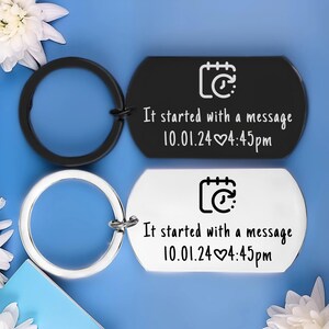 May include: Two keyrings, one black and one silver, each with a tag. The tags are engraved with the text "It started with a message 10.01.24 4:45pm" and a calendar icon. The keyrings are on a blue background.