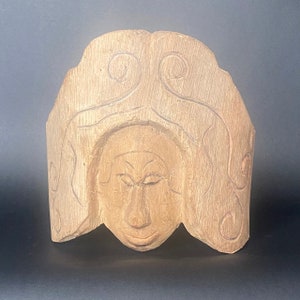 Puede incluir: Una talla de madera de una cara con detalles intrincados. La cara tiene los ojos cerrados y una nariz y boca detalladas. La talla está rodeada por un borde decorativo.