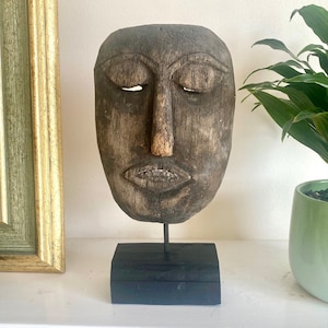 May include: A wooden carved face mask with closed eyes and a neutral expression, mounted on a black wooden stand.