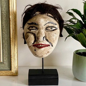 May include: A wooden mask with a painted face, black hair, and a red mouth. The mask is on a black stand.