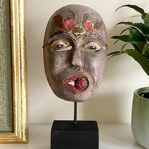 May include: A wooden mask with a painted face and red lips. The mask has a red and gold design on the forehead and is mounted on a black stand.