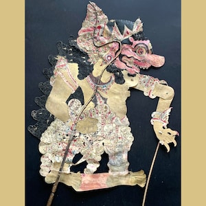 Pode incluir: Um fantoche de sombras tradicional indonésio, ou wayang kulit, que representa uma criatura mítica com uma expressão feroz. O fantoche é feito de couro entalhado e pintado de forma intrincada, com detalhes dourados e vermelhos. O fantoche é segurado por um pau e está pronto para ser usado em uma apresentação de fantoches de sombras.