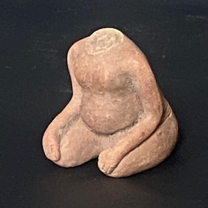 Puede incluir: Pequeña figura sentada sin cabeza, de color marrón claro. Los brazos están doblados y las piernas recogidas. La escultura tiene un vientre redondeado y parece estar hecha de arcilla o un material similar. El fondo es oscuro.