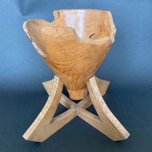 May include: A wooden bowl with a unique, irregular shape, resting on a wooden stand with a geometric design. The bowl is light brown and has a natural wood finish.