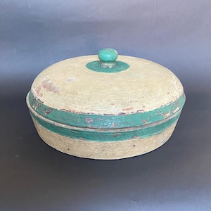 Puede incluir: Una caja redonda de madera con tapa. La caja está pintada de un color beige claro con una banda verde alrededor de la parte superior e inferior. La tapa tiene un pequeño pomo verde en la parte superior.