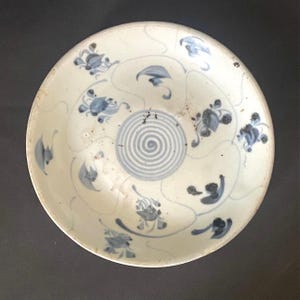 May include: A white ceramic plate with blue floral and leaf patterns. The plate has a central spiral design and a slightly off-white border. The overall design is delicate and detailed.