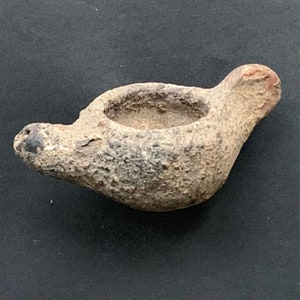 May include: A small, gray, ceramic oil lamp with a spout on one side and a round opening on top. The lamp has a rough, textured surface.