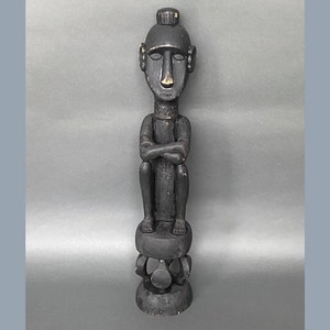 May include: A black wooden sculpture of a seated figure with arms crossed. The figure has a stylized face with a rounded head and a small, round base.