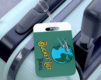 Op de afbeelding: Een turquoise bagagelabel met een witte rand en een wereldbol met een vliegtuig dat eroverheen vliegt. De tekst "Bucket List Travel" is geschreven in een geel handschrift.