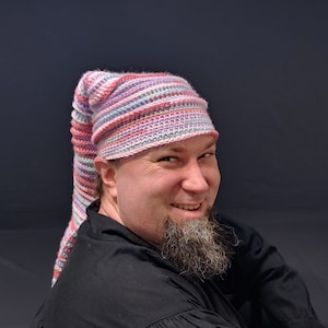 May include: A person wearing a pink, purple, and blue striped crocheted hat with a long tail. The hat is pulled down over their forehead and the person is smiling.