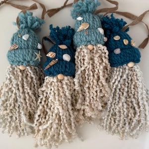 May include: Four gnome ornaments featuring knitted hats in varying shades of blue, each topped with a pom-pom and decorated with small seashells. Each gnome has a round wooden bead nose and a long, textured, cream-colored yarn beard.