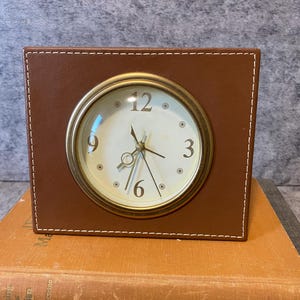 May include: A brown square clock with a gold-coloured circular clock face. The clock has a brown leather-like exterior with white stitching around the edges. The clock face has black hands and the numbers 12, 3, 6, and 9.