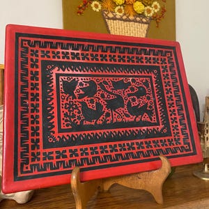May include: A rectangular red and black decorative tray featuring a central panel with black silhouettes of birds and floral motifs, surrounded by a geometric border pattern of repeating black crosses and lines on a red background.