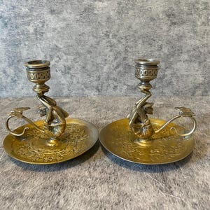 May include: A pair of antique brass candle holders. Each holder features a detailed design with a small figure supporting the candle cup. The holders have a circular base with an ornate floral pattern and a handle.
