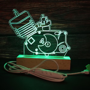Könnte beinhalten: Eine grün beleuchtete Acryl-Lampe in Form eines 4-Takt-Motors mit der Zahl "4" an der Seite. Die Lampe steht auf einem Holzsockel und hat ein weißes Kabel mit einem Schalter.